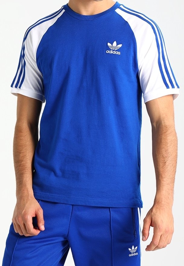 Mies, joka käyttää sinistä Adidas-urheiluasu-settiä, jossa on valkoiset hihnat ja kolme sinistä raitaa hartioilla ja housuissa, seisoo valkoista taustaa vasten.