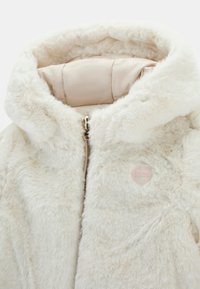 Kremowa kurtka z sztucznego futra z kapturem, złotym zamkiem błyskawicznym, ocieplaną podszewką i małym, sercowym logo Guess na piersi.