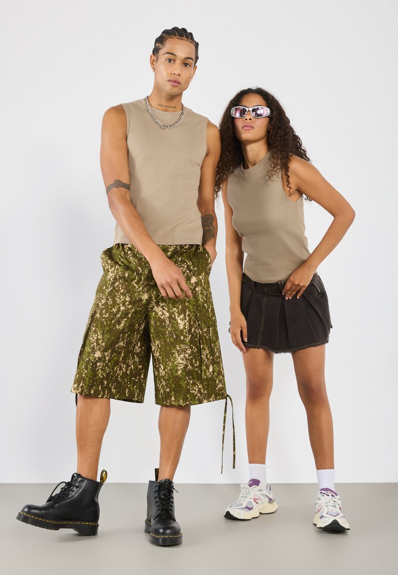 To modeller har koordinerte antrekk. Den mannlige modellen har på seg en beige ermeløs skjorte og grønne camo-shorts, kombinert med svarte støvler. Den kvinnelige modellen bruker en beige ermeløs skjorte og et svart miniskjørt, supplert med flerfargede joggesko og solbriller.