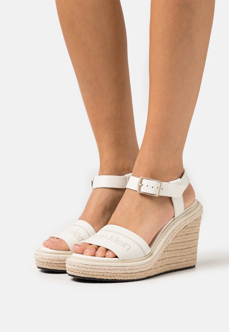 Pieds portant des sandales compensées blanches Calvin Klein avec des brides à la cheville et une plateforme en jute tressé sur un fond uni.