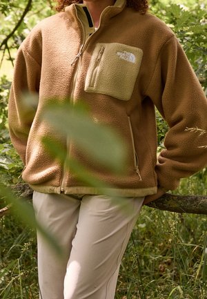 Personne portant une veste polaire beige avec une poche poitrine et un pantalon beige, debout à l'extérieur près de plantes et d'une branche en bois.