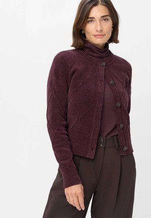 Kvinde iført en mørk lilla strikket cardigan over en matchende højhalset trøje og mørkebrune højttaljede bukser mod en ensfarvet baggrund.