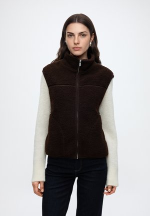 Vrouw draagt een donkerbruine fleece bodywarmer zonder mouwen over een witte trui met lange mouwen en donkere broek, staande tegen een effen achtergrond.