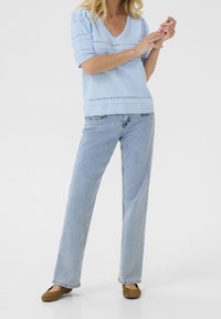 Mujer vestida con un suéter de punto en azul claro con cuello en V, jeans rectos en azul claro y zapatos planos marrones, de pie sobre un fondo blanco.