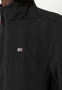 Svart jacka med hög krage, prydd med en liten flerfärgad logotyp. Tyget har en slät yta utan synliga mönster.