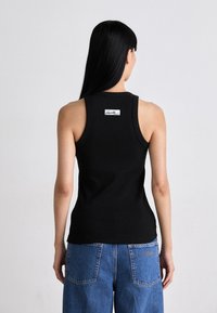 Frau mit langen schwarzen Haaren, die ein schwarzes ärmelloses Oberteil mit einem kleinen weißen Logo auf dem oberen Rücken und blaue Jeans trägt.