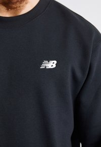 Czarna bluza z bawełny z dodatkiem włókien syntetycznych, z okrągłym dekoltem. Posiada małe, białe logo "NB" na lewej piersi. Gładka tekstura.