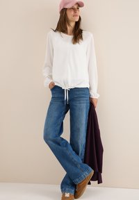 Blusa blanca de manga larga con cordón en la cintura, combinada con jeans de campana azules y zapatillas marrones, complementada con una gorra rosa.