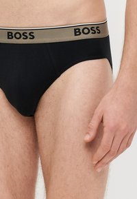 Juodi medvilniniai apatiniai su auksiniu ir juodu elastiniu juosmeniu, ant kurio matomas "BOSS" logotipas. Artimas vaizdas pabrėžia formą ir audinio tekstūrą.