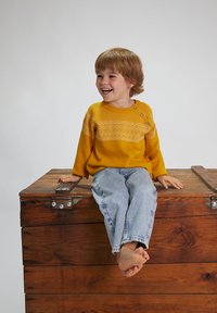 Maglione giallo lavorato a maglia con un motivo geometrico e tre bottoni, abbinato a jeans di denim azzurro chiaro. Bambino seduto su una cassa di legno.