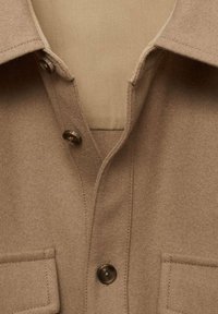 Beige wolblend overhemd met een puntige kraag, twee voorkruiszakken en donkere knopen; gladde textuur en zichtbare stikseldetails.