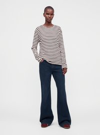 Femme portant une chemise à manches longues blanche et noire rayée avec un jean large bleu foncé et des chaussures marron, debout avec une main dans la poche.