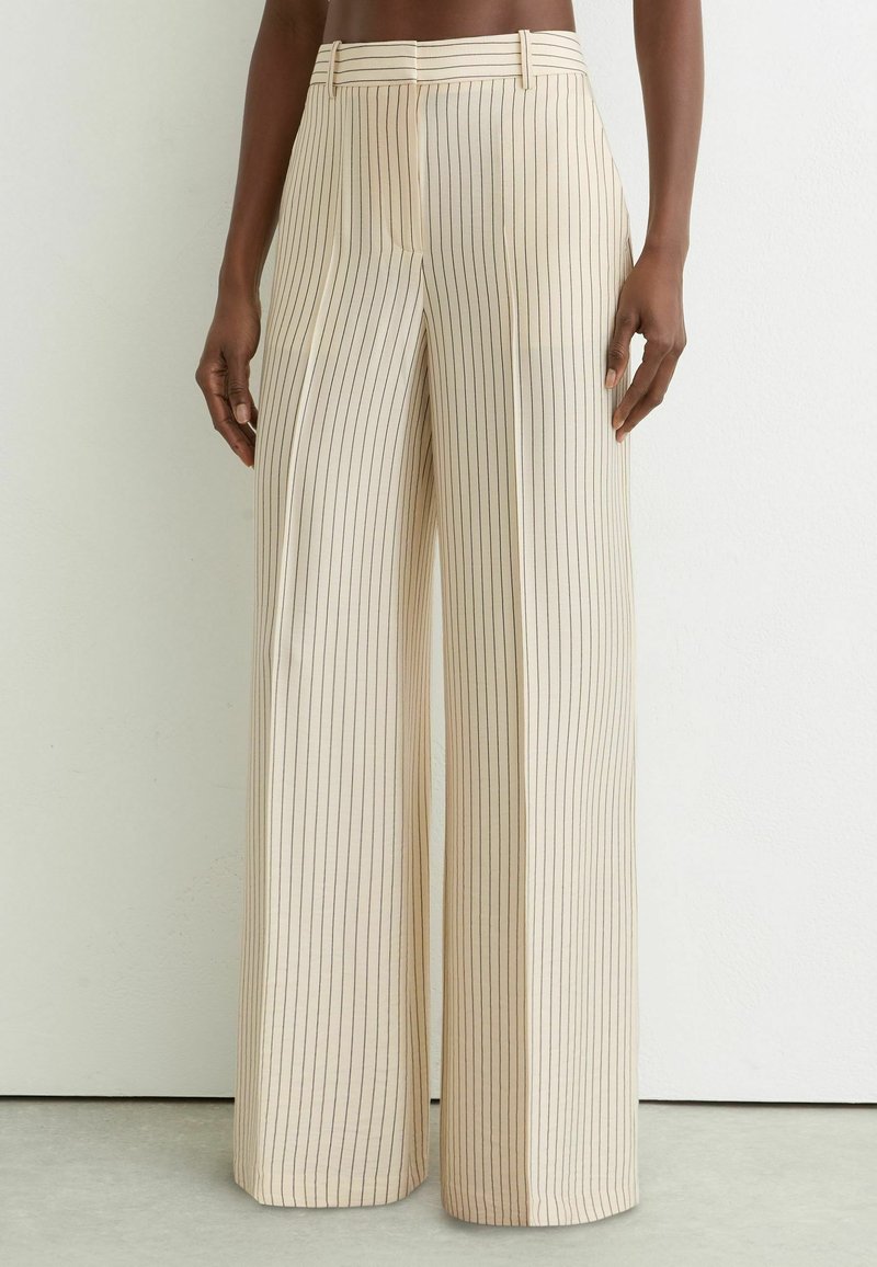 Pantalones beige de pierna ancha con finas rayas verticales negras, cintura alta, presillas para cinturón, usados por una persona que está de pie contra una pared blanca lisa.