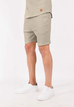 Mann in beigen, strukturierten Shorts und passendem Hemd mit weißen Sneakers auf weißem Hintergrund, steht mit sichtbaren Beinen.