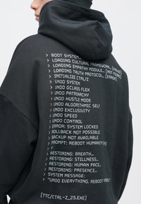 Μαύρη φούτερ με κουκούλα, που έχει λευκό εκτυπωμένο κείμενο που μοιάζει με κώδικα υπολογιστή στο πίσω μέρος. Το κείμενο περιλαμβάνει εντολές και μηνύματα σφάλματος.