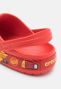 Red plastic Crocs featuring a smooth upper, two wide straps, and a design with a cartoon car and flames on the side. Text "Rust-eze" included.