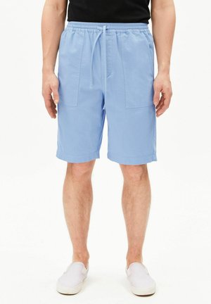 Mann trägt hellblaue Shorts mit Kordelzug und weiße Slip-on-Schuhe, steht vor einem weißen Hintergrund.