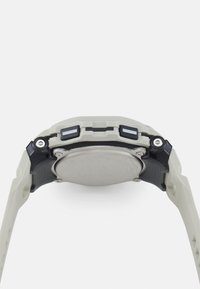 Montre gris clair avec un boîtier rond en métal et des éléments en noir. Elle possède un bracelet texturé, deux boutons latéraux et un dos circulaire lisse.