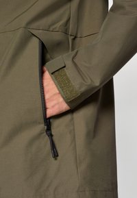 Χακί πράσινο μπουφάν με τσέπη φερμουάρ και μανσέτα με βέλκρο. Λείο, ελαφρύ υλικό με ματ υφή. Ρυθμιζόμενη κορδόνι στο κάτω μέρος.