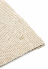 Żebrowany beżowy dodatek z wełny z małą złotą metką. Tekstura jest miękka i ciepła, podkreślająca jego dzianinowy design i rzemiosło.