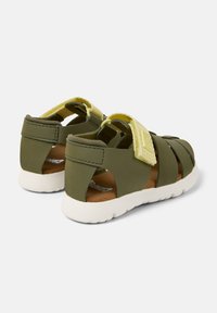 Två gröna sandaler med stängd bak och öppen tå, som har justerbara gula remmar, texturerade ytor och en vit gummisula.
