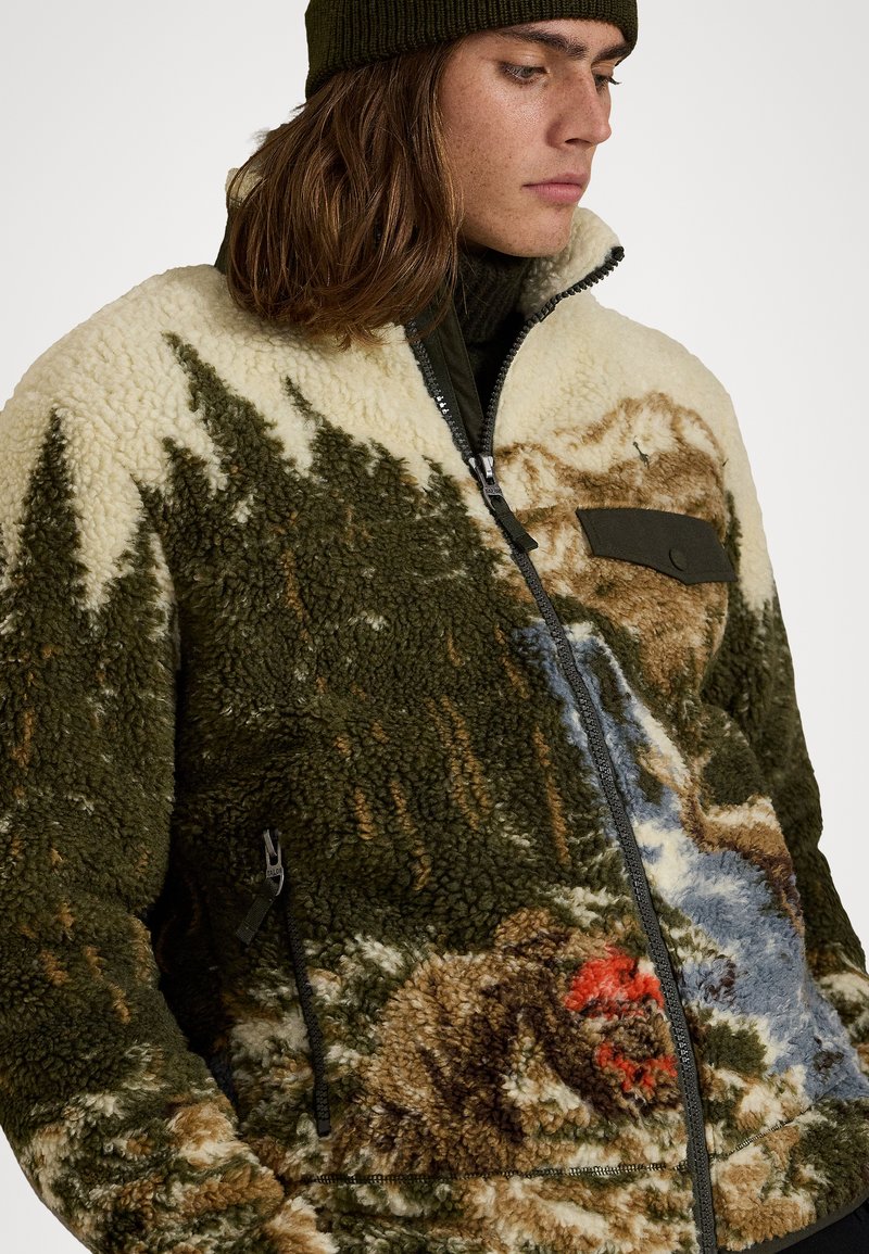 Fleecejacka med ett naturinspirerat mönster som innehåller träd, kullar och en flod. Färgschema i beige, grönt och brunt. Dragkedja fram.