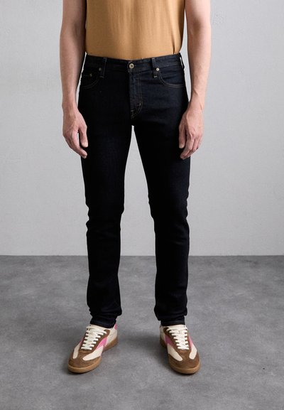 Jeans in denim scuro con vestibilità slim, dotati di cinque tasche e cuciture gialle a contrasto. Abbinati a scarpe da ginnastica multicolori.