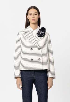 White and black striped double-breasted jacket with large collar and decorative navy flower accent, worn over dark denim pants.