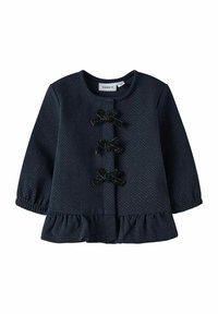 Top de manga larga color navy con lunares blancos, dobladillo con volantes, escote redondo y tres lazos decorativos negros en la parte delantera. Textura suave.