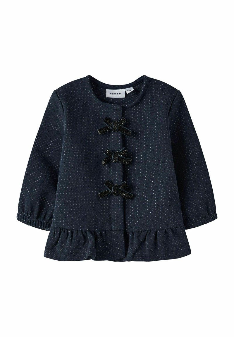 Top de manga larga color navy con lunares blancos, dobladillo con volantes, escote redondo y tres lazos decorativos negros en la parte delantera. Textura suave.