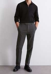 Pullover nero con zip e maniche lunghe abbinato a pantaloni grigi su misura. I pantaloni presentano una texture sottile e un taglio dritto. Scarpe nere con lacci.