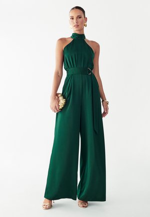 Kvinde iført en ærmeløs, emeraldgrøn halter-jumpsuit med vide ben, guldbøjler, armbånd, remsandaler og holder en guldtaske.