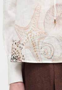 Λευκό ύφασμα με αστερίες σε μπεζ και καφέ αποχρώσεις, κοχύλια και σχέδιο με στίγματα, φορεμένο πάνω από καφέ υφασμάτινο παντελόνι, με εμφανές χέρι στο πλάι.