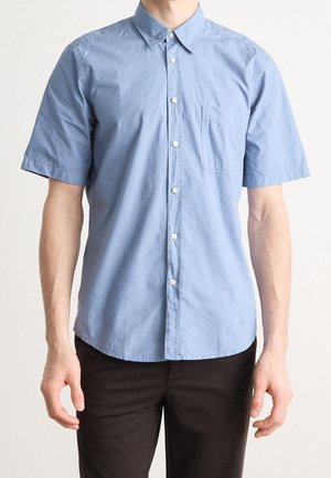 Mężczyzna w jasnoniebieskiej koszuli na krótki rękaw z kieszenią na piersi i czarnych spodniach, stojący na tle jednolitego jasnego tła.