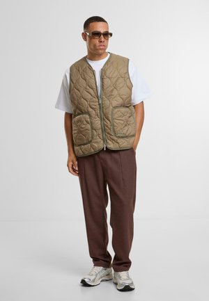 Padded beige vest with green trim, featuring a zip front and two large front pockets. Worn over a white shirt and paired with brown trousers.