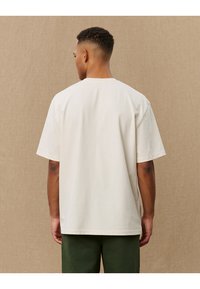 Cremfarbener, weicher Baumwoll-T-Shirt mit kurzen Ärmeln und lockerer Passform, von hinten betrachtet. Glatte Textur, keine sichtbaren Muster.