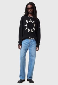 Pulover negru cu text circular alb "SAINT", asortat cu blugi largi de culoare albastră deschisă. Modelul poartă ochelari de soare negri și încălțăminte neagră.