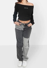 Musta ribbed off-the-shoulder paita valkoisella logolla, yhdistettynä harmaisiin patchwork-farkkuihin ja valkoisiin tennareihin. Keskittyminen rentoon katutyyliseen muotiin.