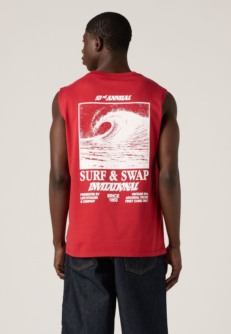 Mann trägt rotes ärmelloses Shirt mit weißem Surf-Wellen-Grafik und dem Text "Surf & Swap Invitational" auf dem Rücken.