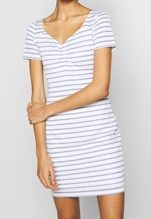 Kobieta ubrana w białą sukienkę z krótkim rękawem, ozdobioną poziomymi niebieskimi paskami i dekoltem w serek, stojąca na tle jednolitego jasnego tła.