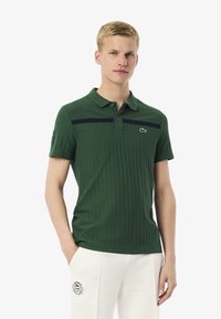 Mörkgrön polo skjorta med ribbad textur, marinblå horisontell rand och broderad logotyp på vänster bröst. Korta ärmar och kragsdesign.