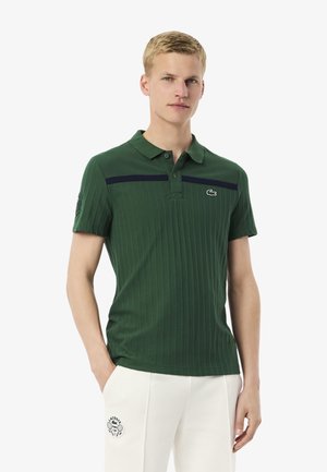 Polo de color verde oscuro con textura acanalada, franja horizontal en color marino, y logo bordado en el lado izquierdo del pecho. Diseñado con mangas cortas y cuello.