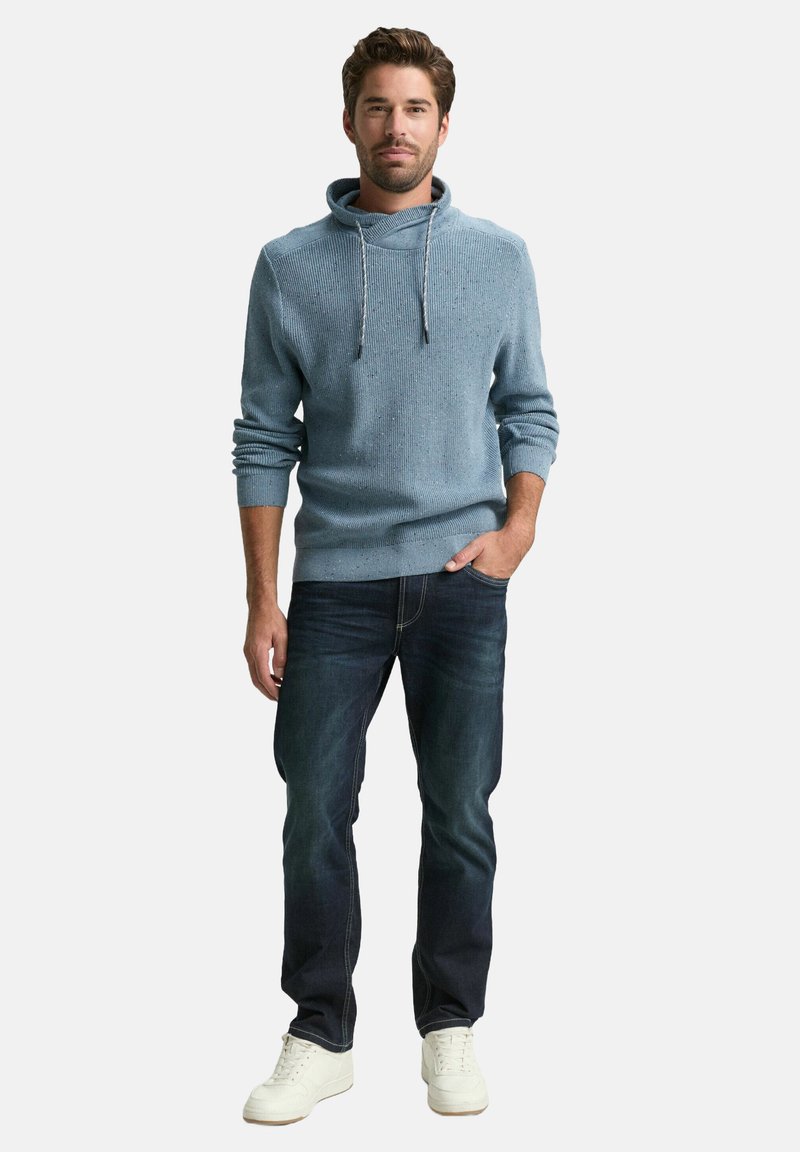 Hellblauer Strickpullover mit Kapuze und Kordelzug, kombiniert mit dunklen Jeans und weißen Sneakern. Lässig geschnitten.