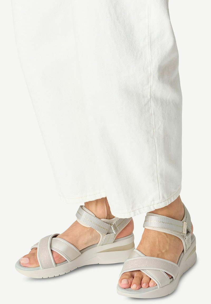 Sandales grises claires avec des brides croisées, des brides de cheville réglables, des accents texturés et une semelle en plate-forme compensée. Portées avec un pantalon blanc.