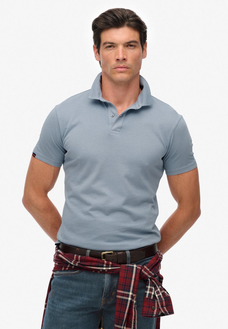 Jasnoniebieska koszulka polo z złożonym kołnierzykiem, uzupełniona wstążką w kratkę z flaneli noszoną w pasie. Dżinsy i czarny pasek dopełniają całość.