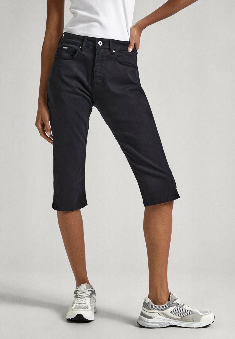 Persona vestida con pantalones capri negros, camiseta sin mangas blanca y zapatillas blancas-grises, de pie con una mano en la cadera frente a un fondo plano.