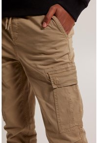 Khaki cargo-housut, joissa on teksturoitu kangas, sivutaskut ja hillityt ommeltyöt. Pikkutaskussa on näkyvissä brodeerattu logo.