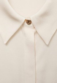 Gros plan d'une chemise crème montrant un col pointu avec un seul bouton décoratif doré au niveau du cou.