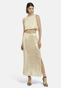 Gelaagd tweedelig outfit in lichtbeige, met een cropped top en een lange rok met een zijsplit. Soepele textuur en glinsterende afwerking.