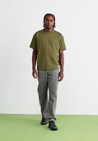 Olivengrønn t-skjorte med kort erm og avslappet passform, sammen med grå rette jeans og sorte joggesko på et lys grønt gulv.